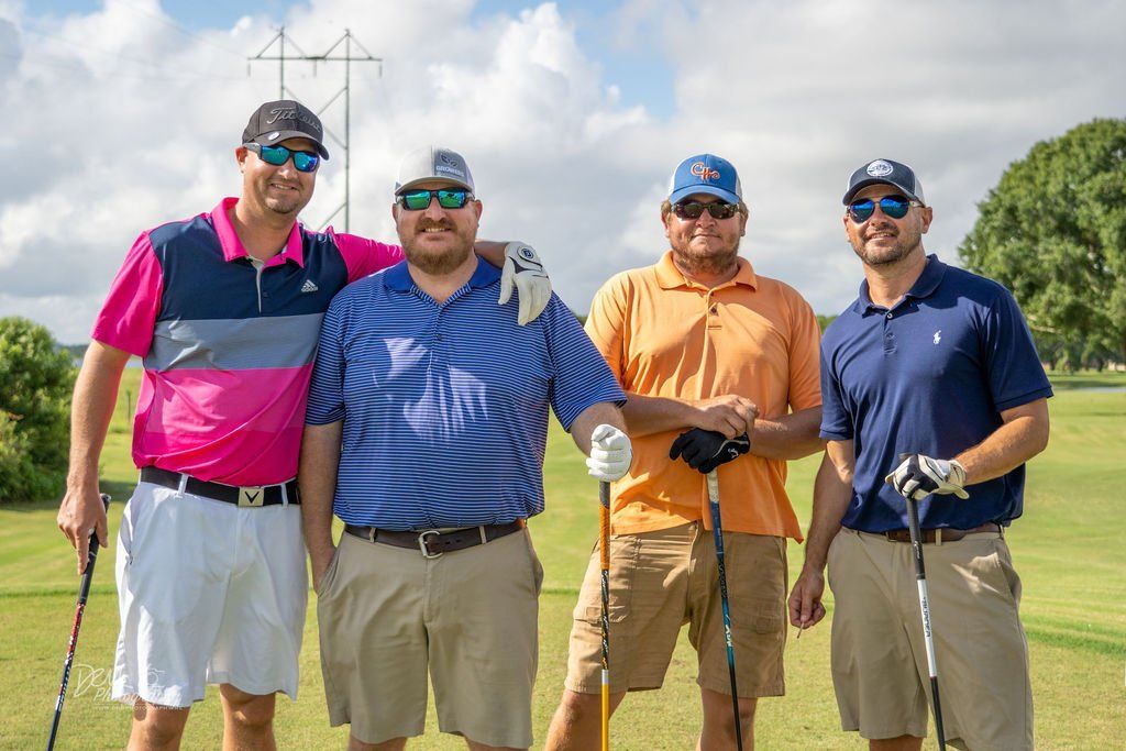 Four men smiling for a photo on a golf course. They wear golf attire and sunglasses, with clubs.