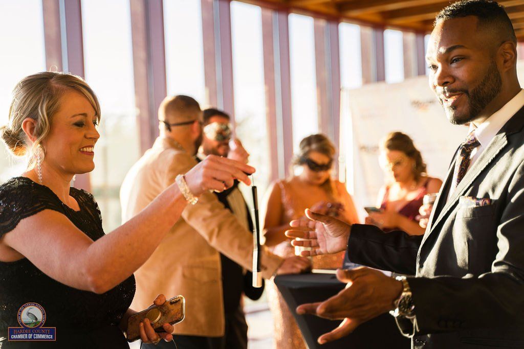 A man in a suit performs a magic trick for a smiling woman. They are indoors near a window, with other people watching.
