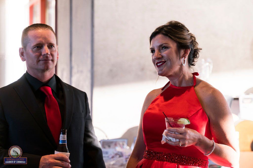 Man in black suit with red tie and woman in red dress at a party. She holds a drink, smiling.