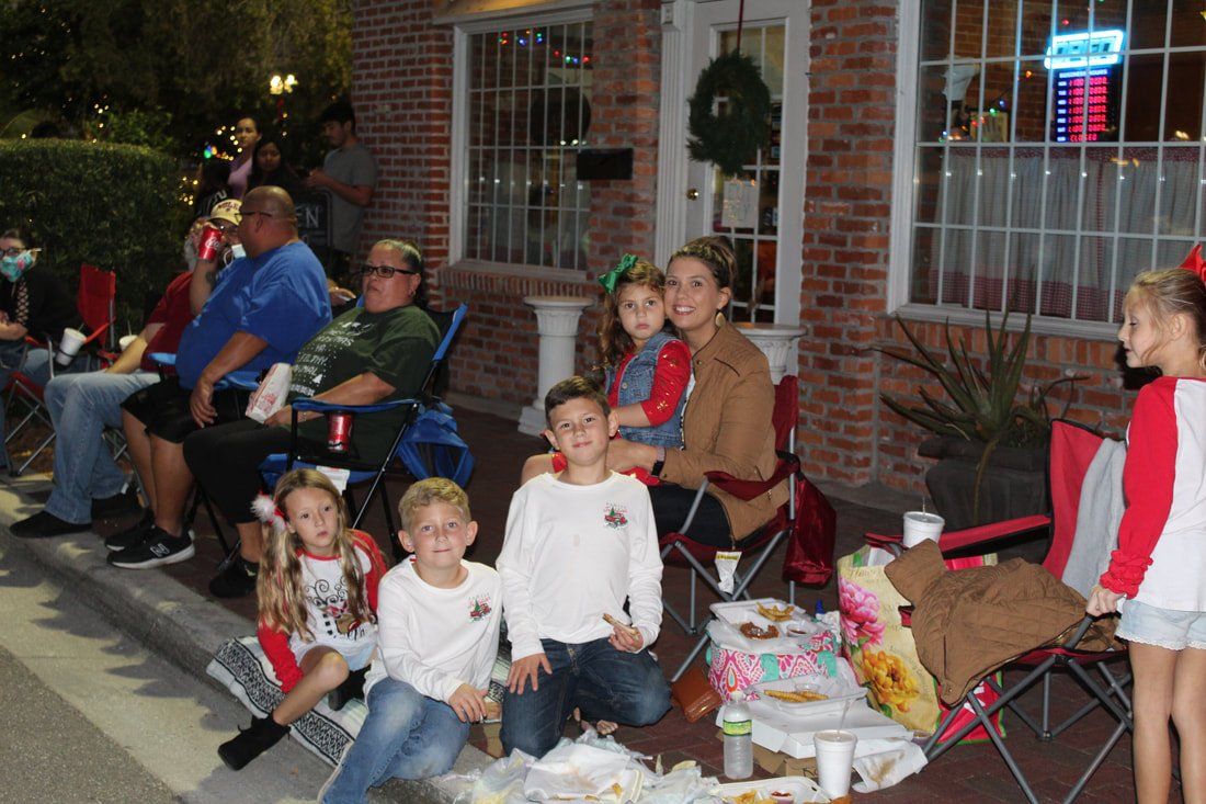 People sitting in chairs, waiting for a parade, near a building decorated for Christmas; a small group of children smiles at the camera.