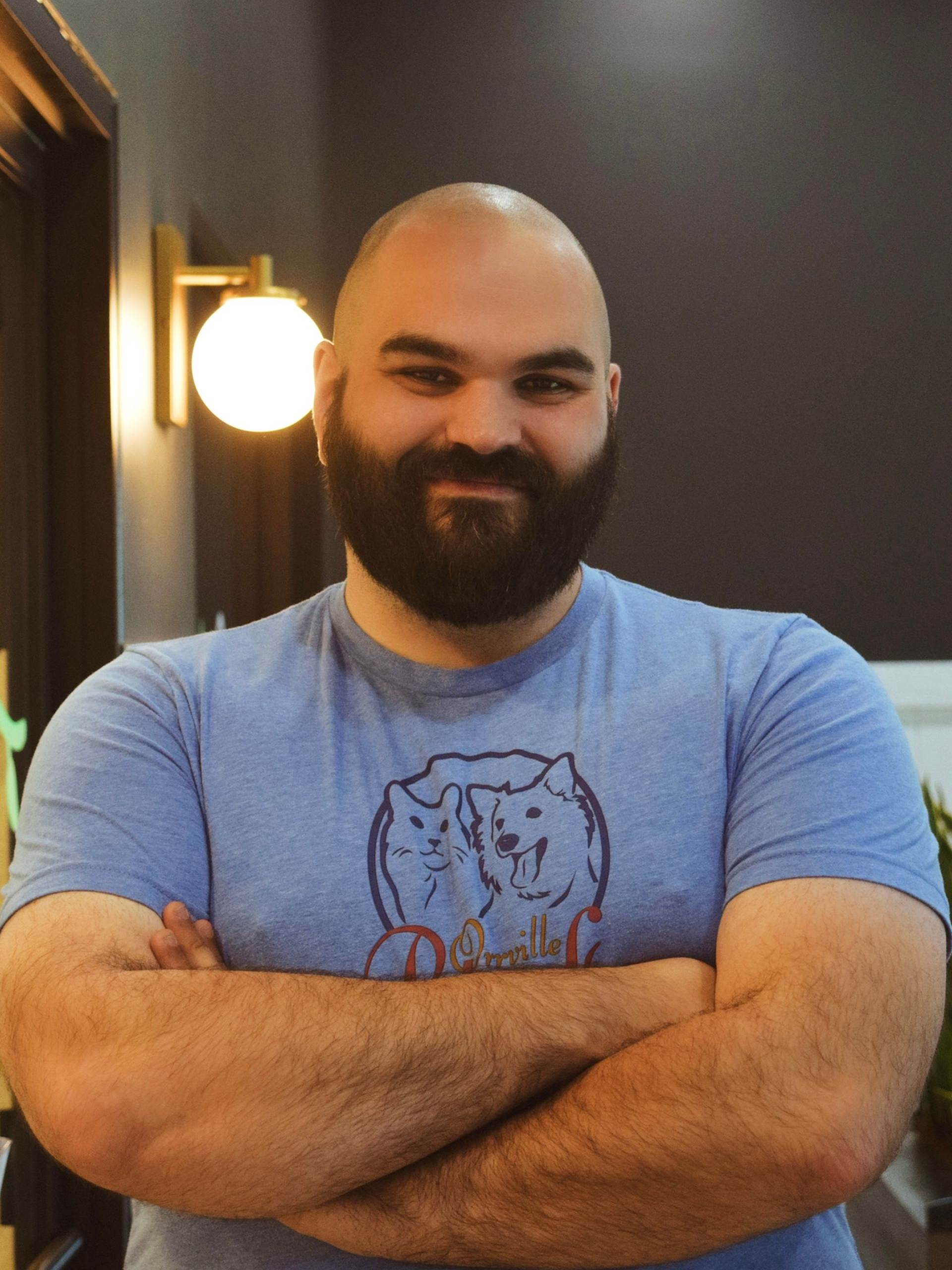 Smiling man with a beard wearing a blue t-shirt, standing inside a building with a modern design, and a light fixture visible.