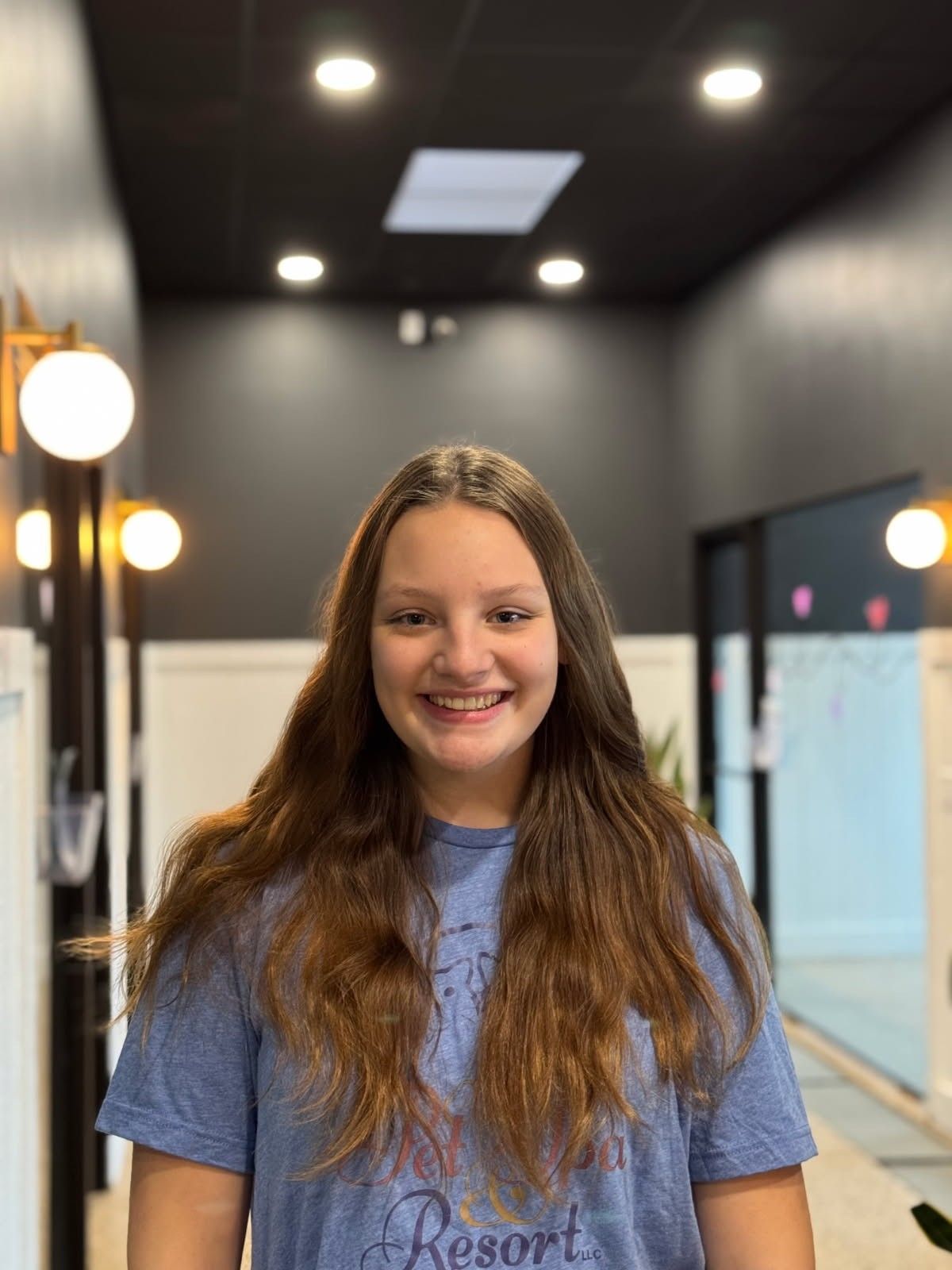 Young woman with long blonde hair sitting in a brown chair, wearing a blue t-shirt, smiling. She is in a modern interior with plants.