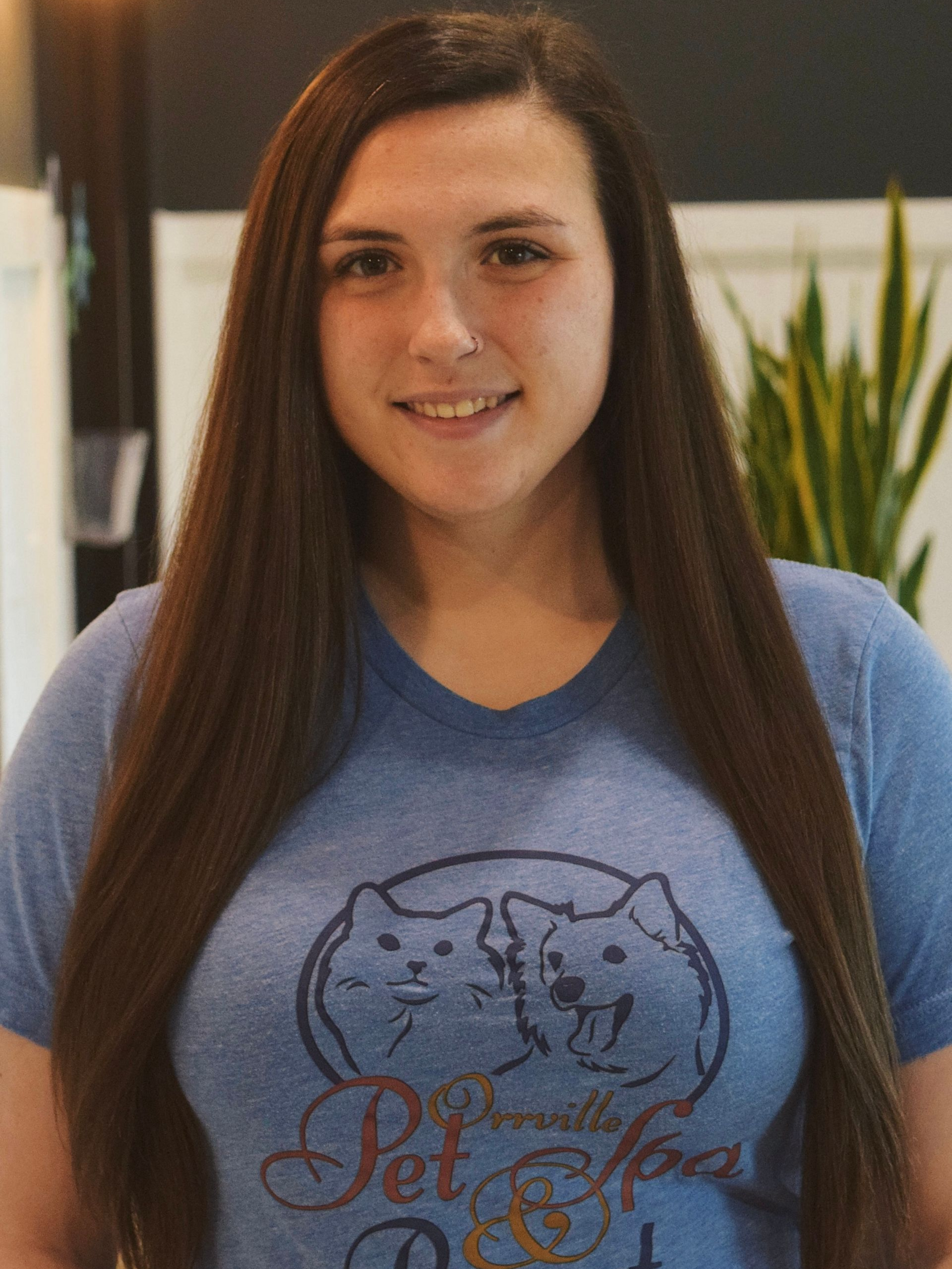 Young woman with long brown hair in a blue shirt, smiling and looking at the camera. Standing in a room with light-colored walls.