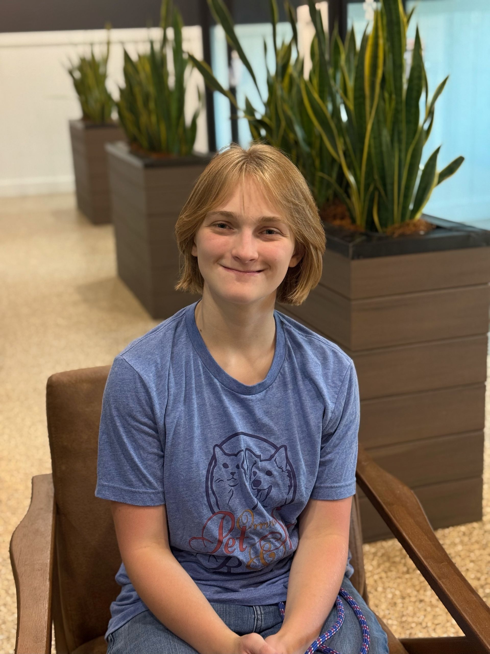 Young person with short, light brown hair, smiling, sitting in a brown chair in front of potted snake plants.