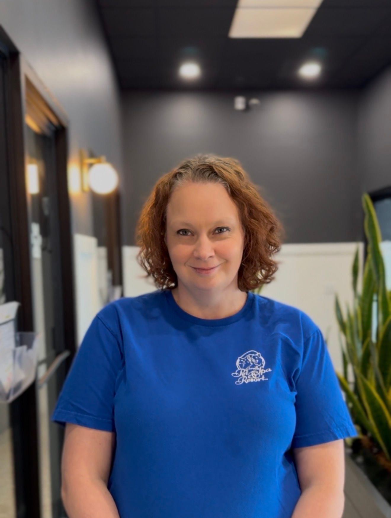 Woman with curly hair in blue shirt and jeans, seated in a chair, smiling. She's in a room with plants and dark accents.
