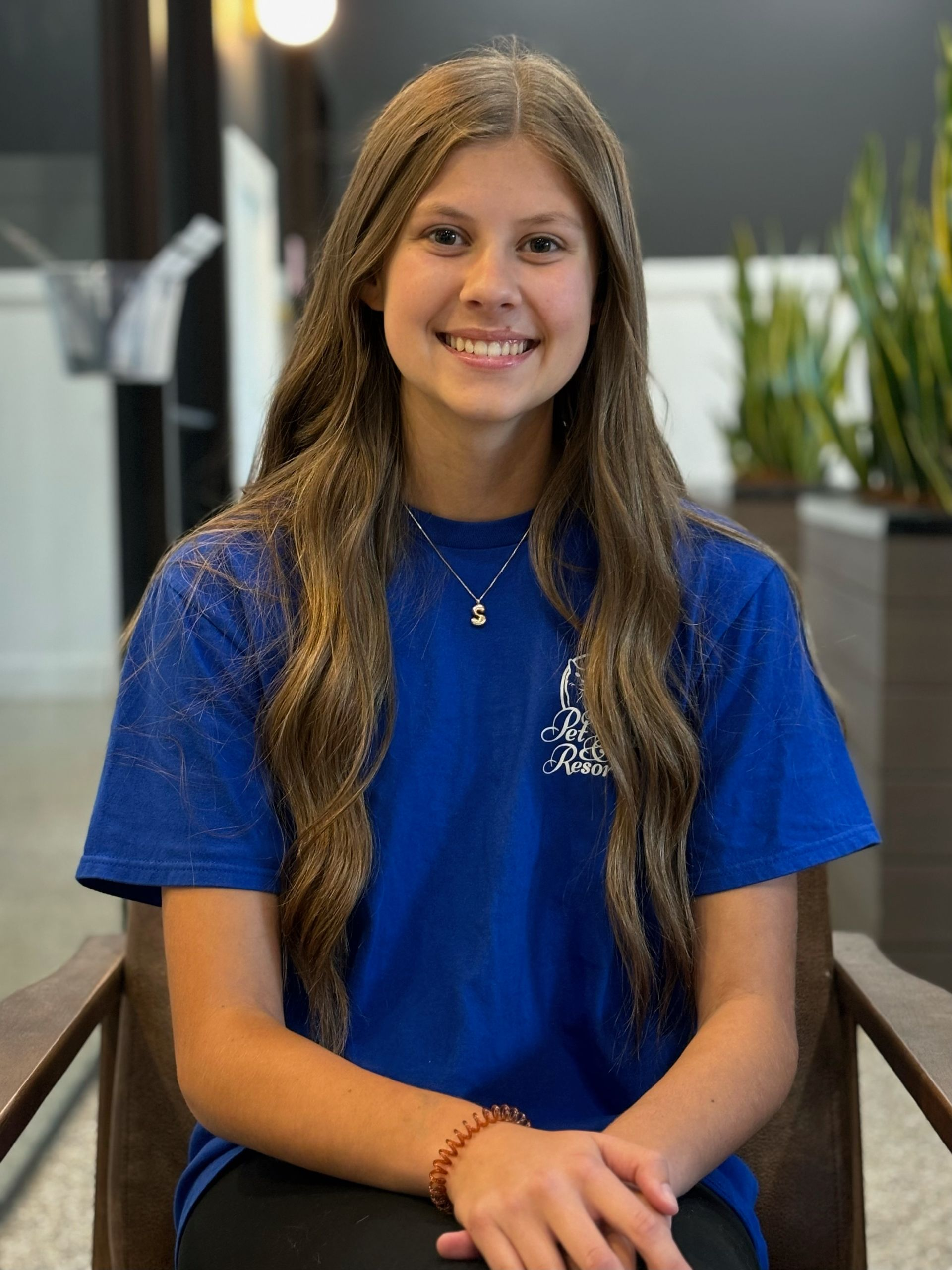 Young woman with long brown hair smiles, wearing a blue shirt, seated indoors.