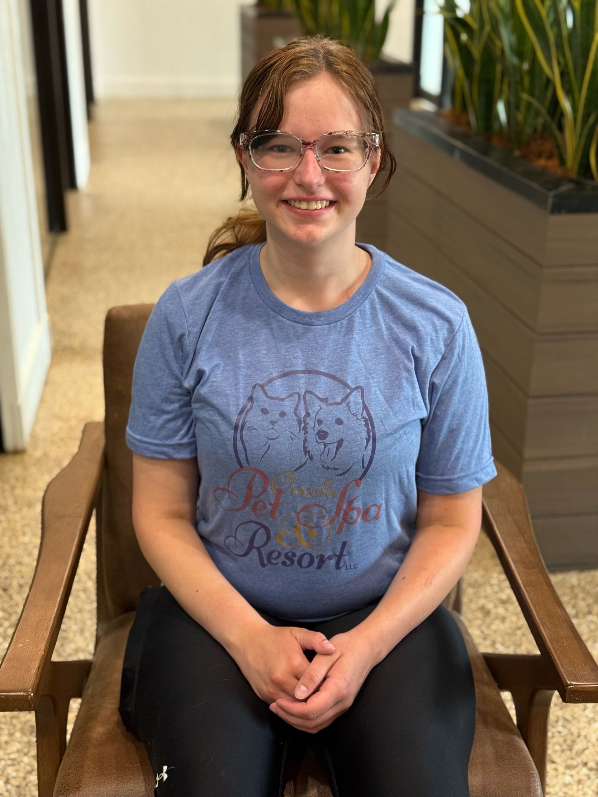 Woman with glasses sits in a wooden chair, wearing a blue shirt with a wolf design and black leggings, indoors. She is smiling.