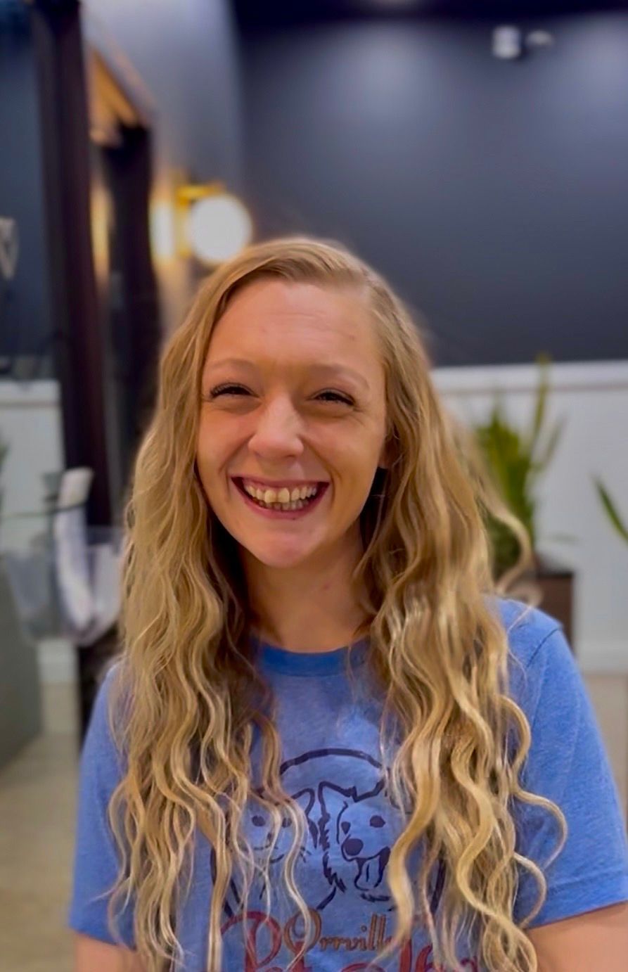 Blonde woman smiling in a blue t-shirt and jeans, sitting in a brown leather chair, in a modern office setting.