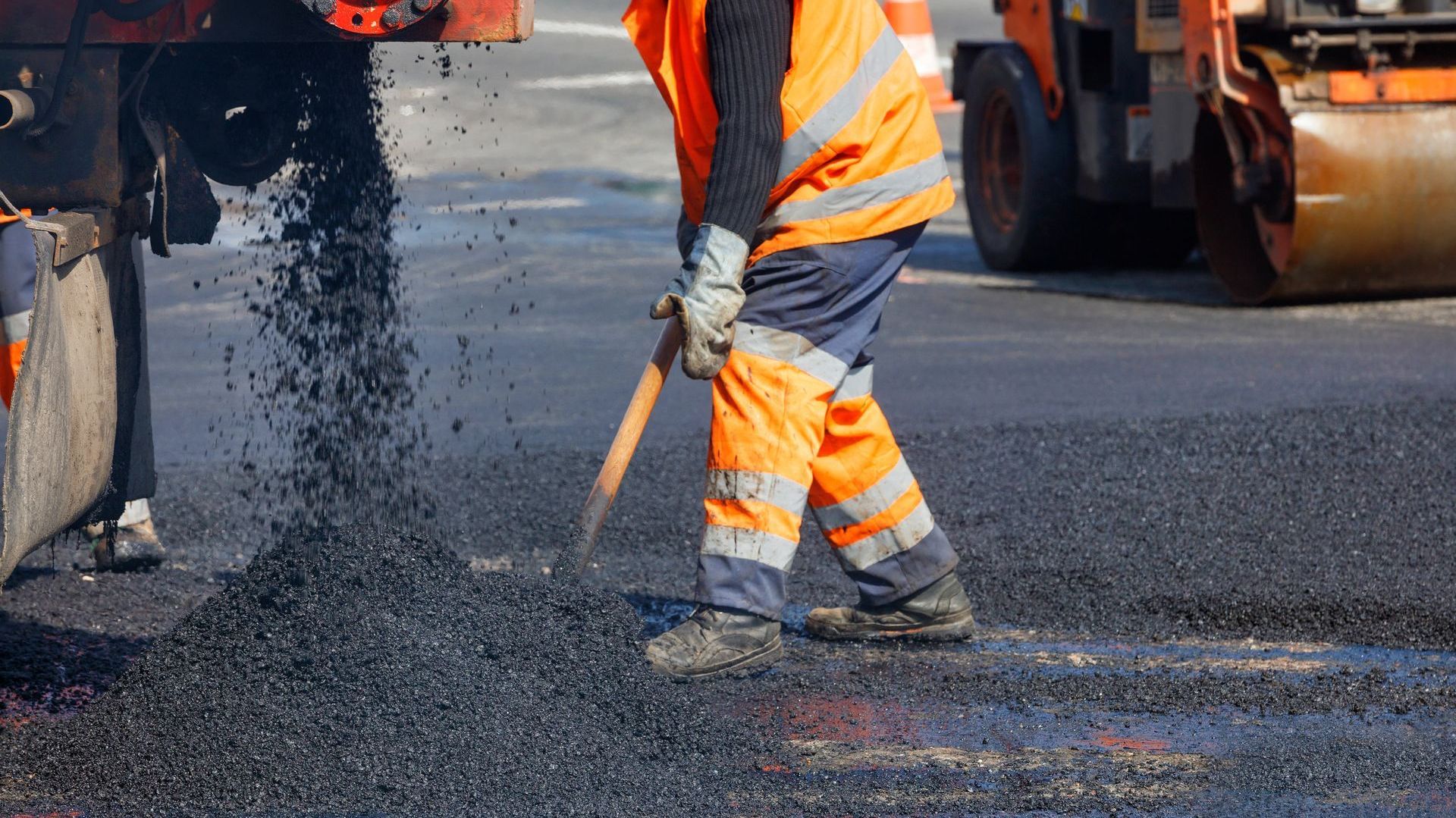 A construction worker in an orange vest pours and levels asphalt, he uses methyl soyate to clean up after
