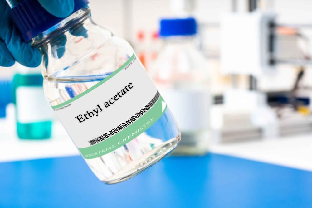 A hand holding a bottle of Ethyl acetate in a laboratory setting.