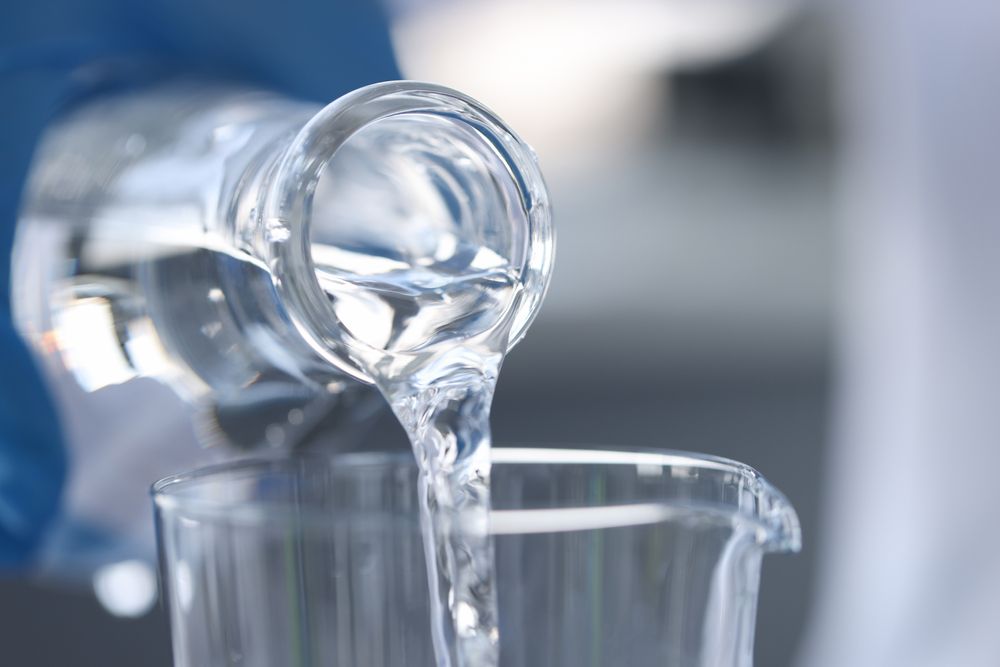 Clear liquid being poured from a glass bottle into a beaker, likely in a lab setting.