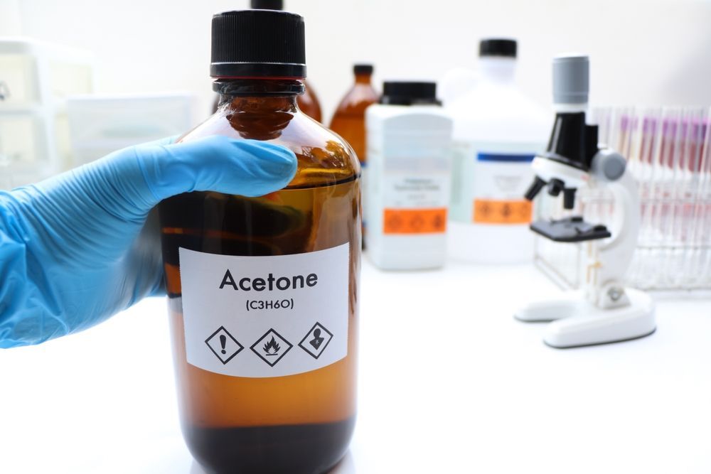 A gloved hand holds a brown bottle labeled Acetone in a laboratory setting with a microscope in the background.