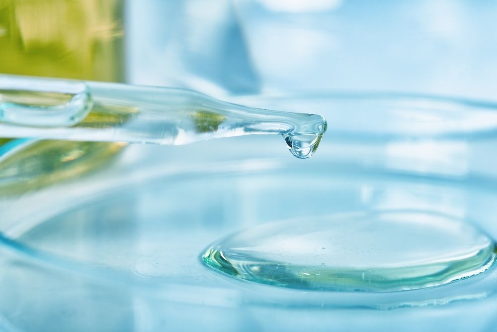 Droplet of clear liquid about to fall from a pipette into a glass container; light blue background.