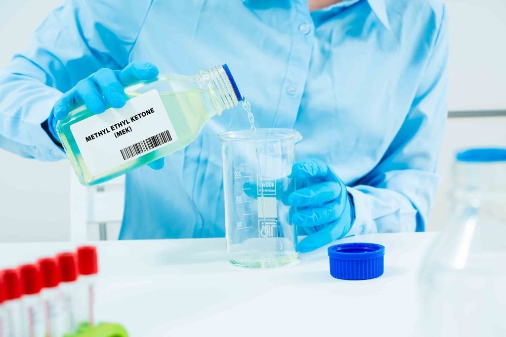 A person in a lab coat and blue gloves pours a clear liquid from a labeled bottle into a glass beaker on a table.