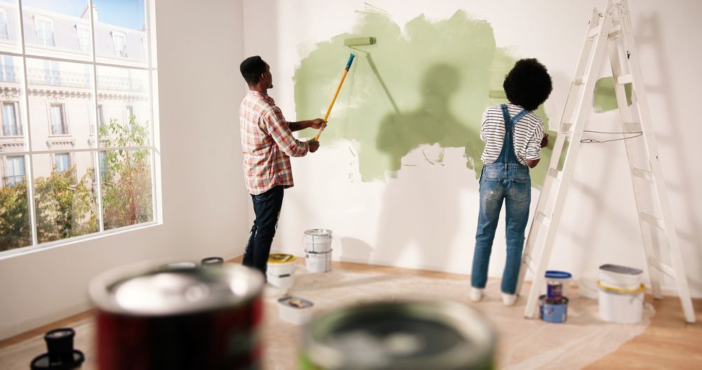 an African American couple, a man in a red flannel and a woman in overalls, paint their wall green while wondering 'what is paint VOCs and are they dangerous'