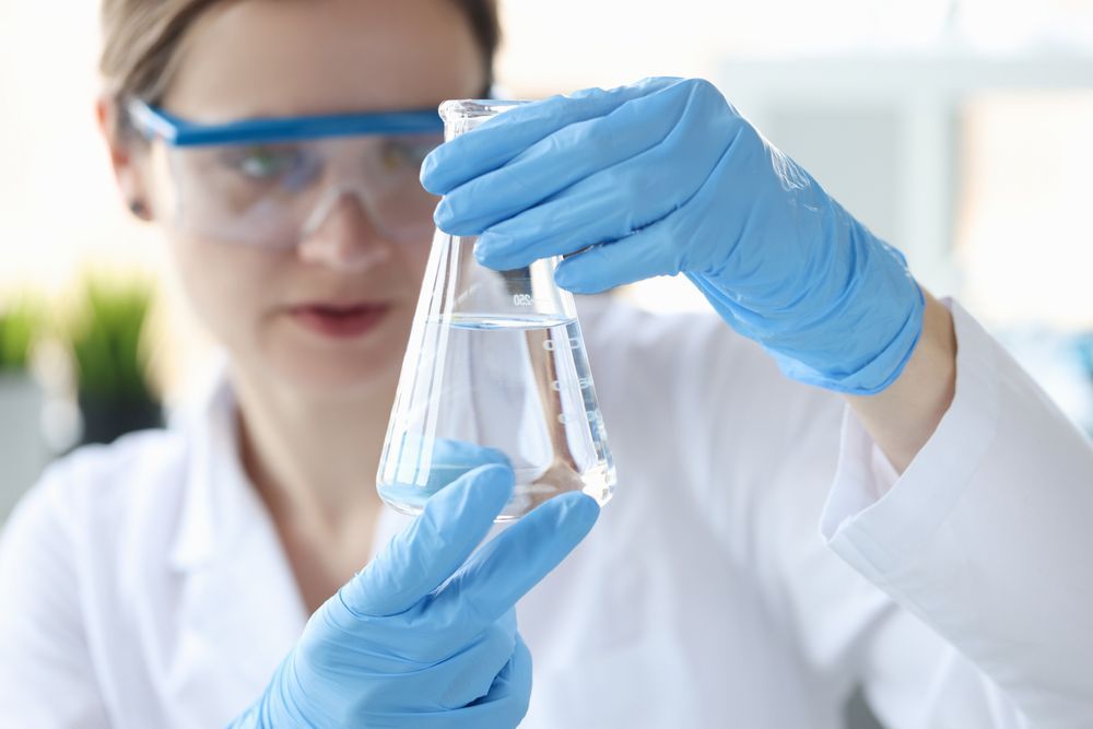a woman in a lab coat and blue gloves is holding a beaker of solvent