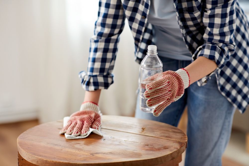 A woman is cleaning a wooden table with a cloth and a bottle of water.