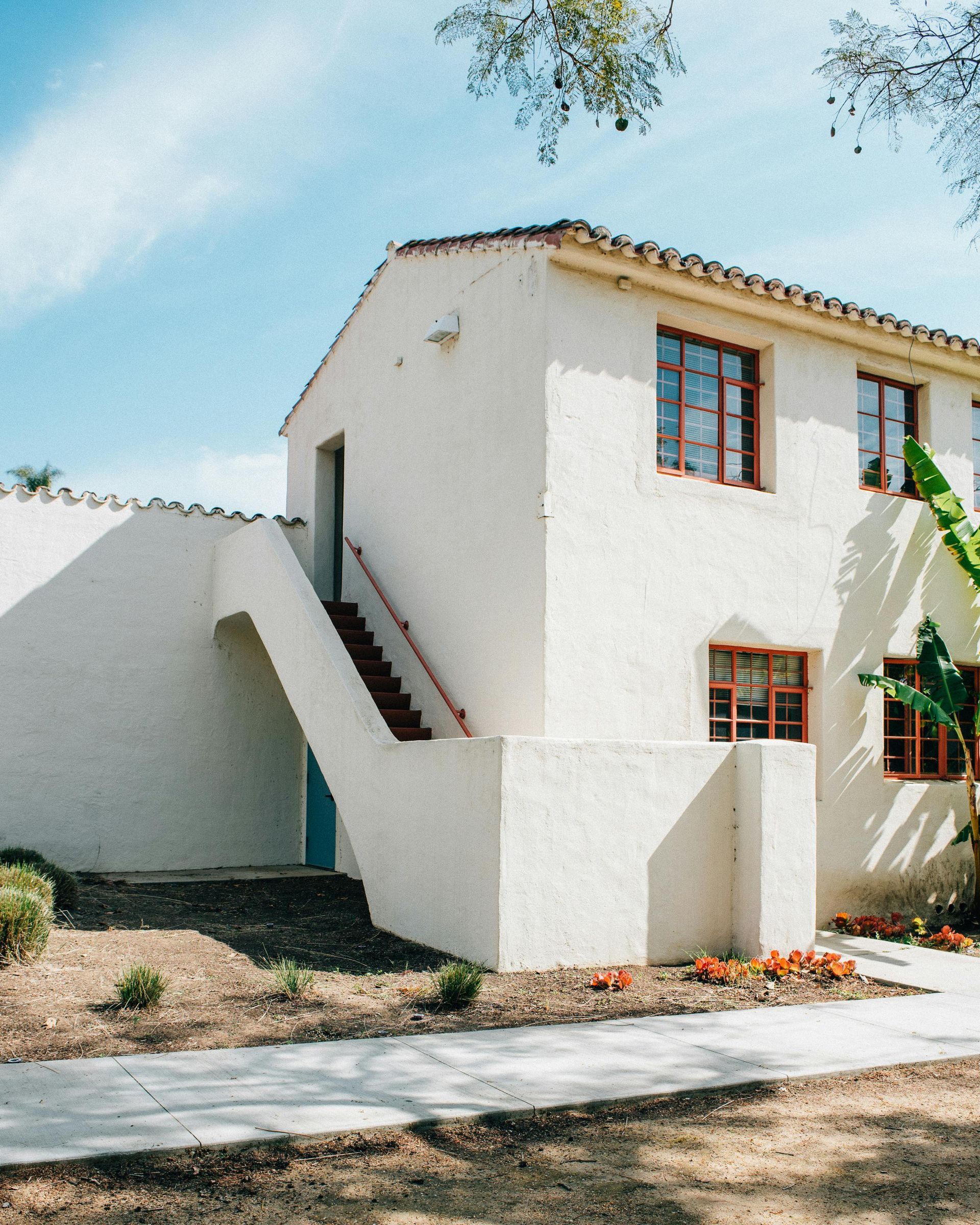 White stucco building with brown-framed windows, red staircase, and blue door. Sunny day.