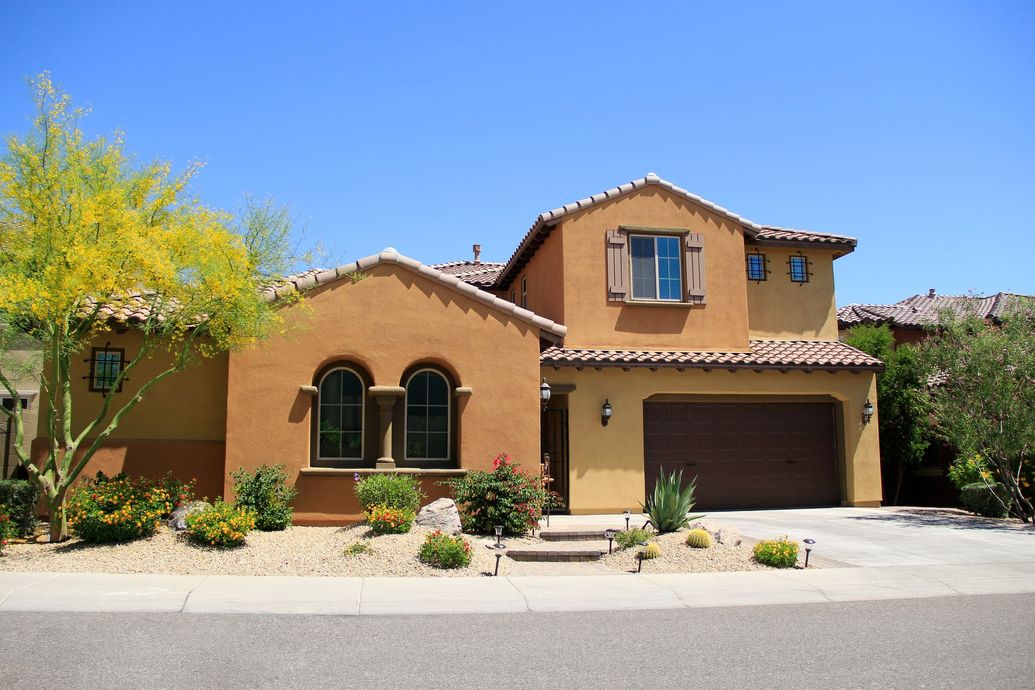 Two-story stucco house with a brown garage door, arched windows, and a tiled roof, under a blue sky.