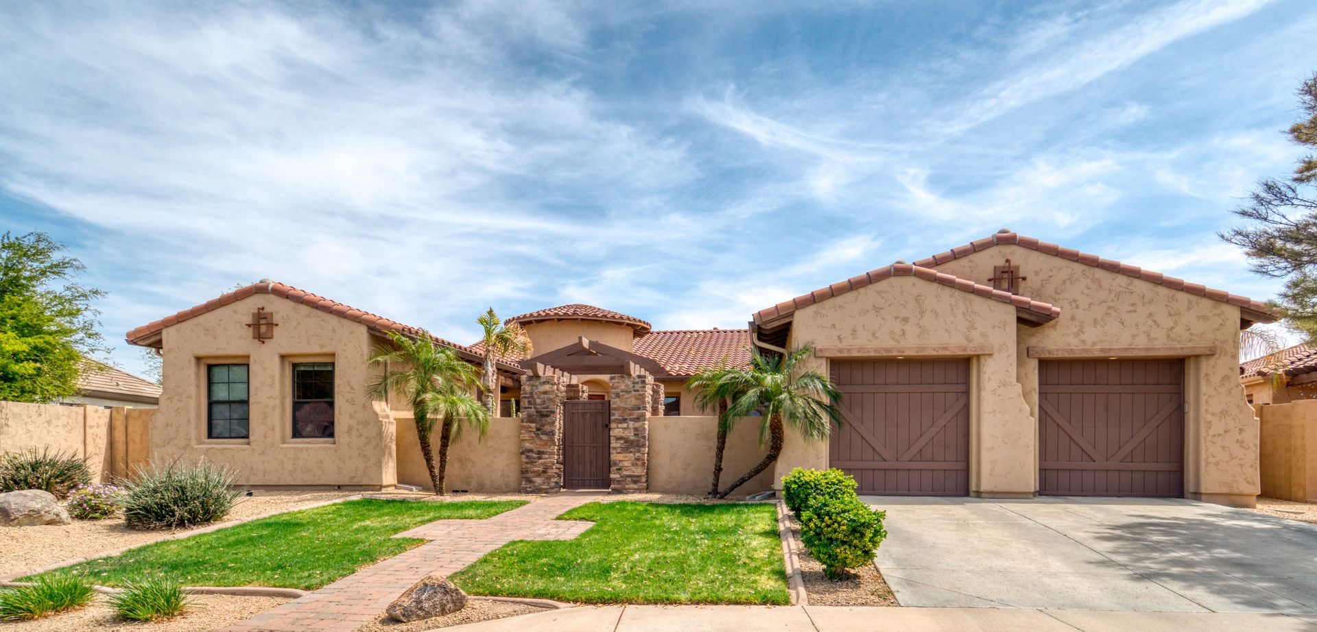 Beige stucco house with a tiled roof, arched entrance, and a two-car garage under a cloudy blue sky.