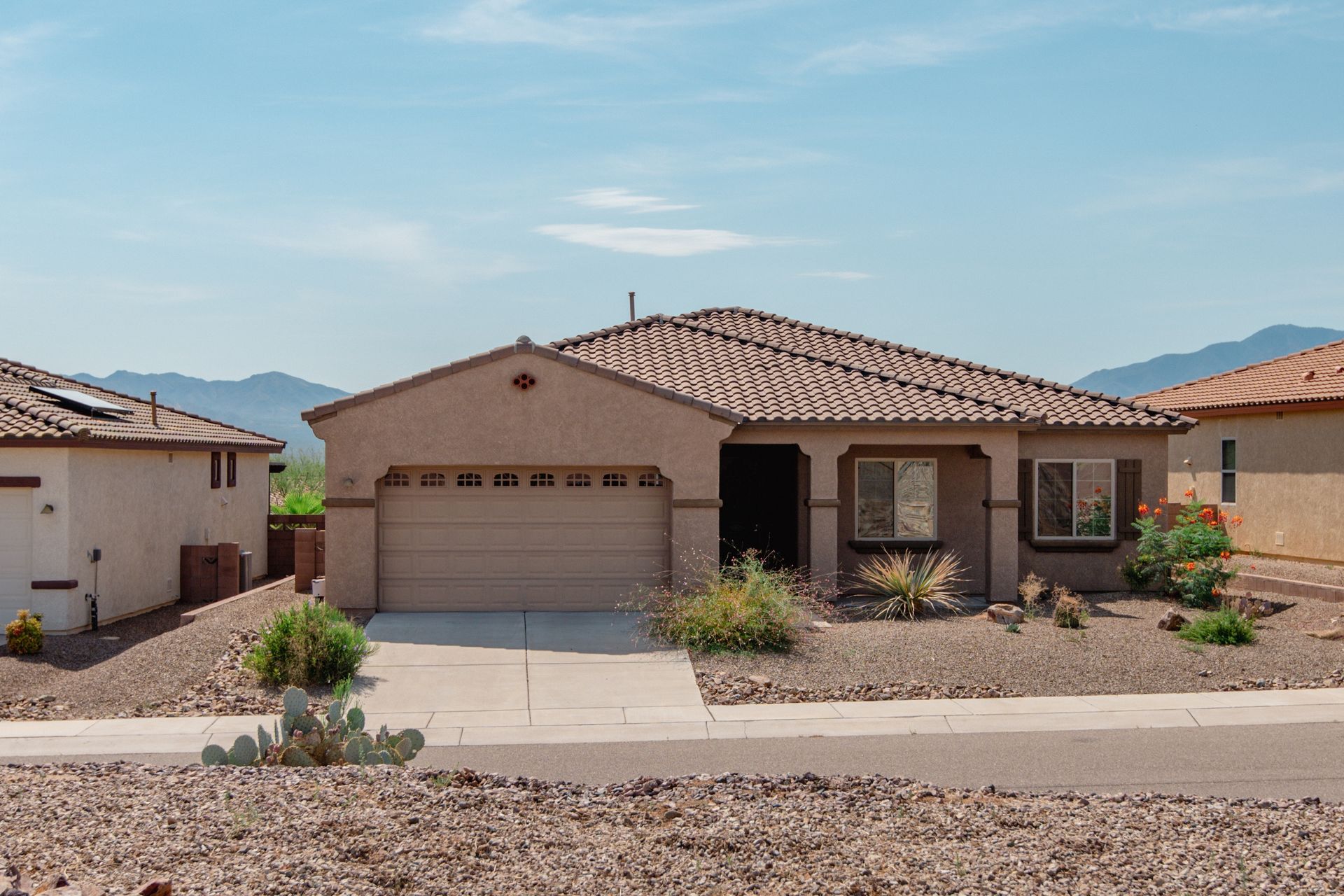 Tan one-story house with a terracotta roof, driveway, and sparse landscaping against a mountain backdrop.