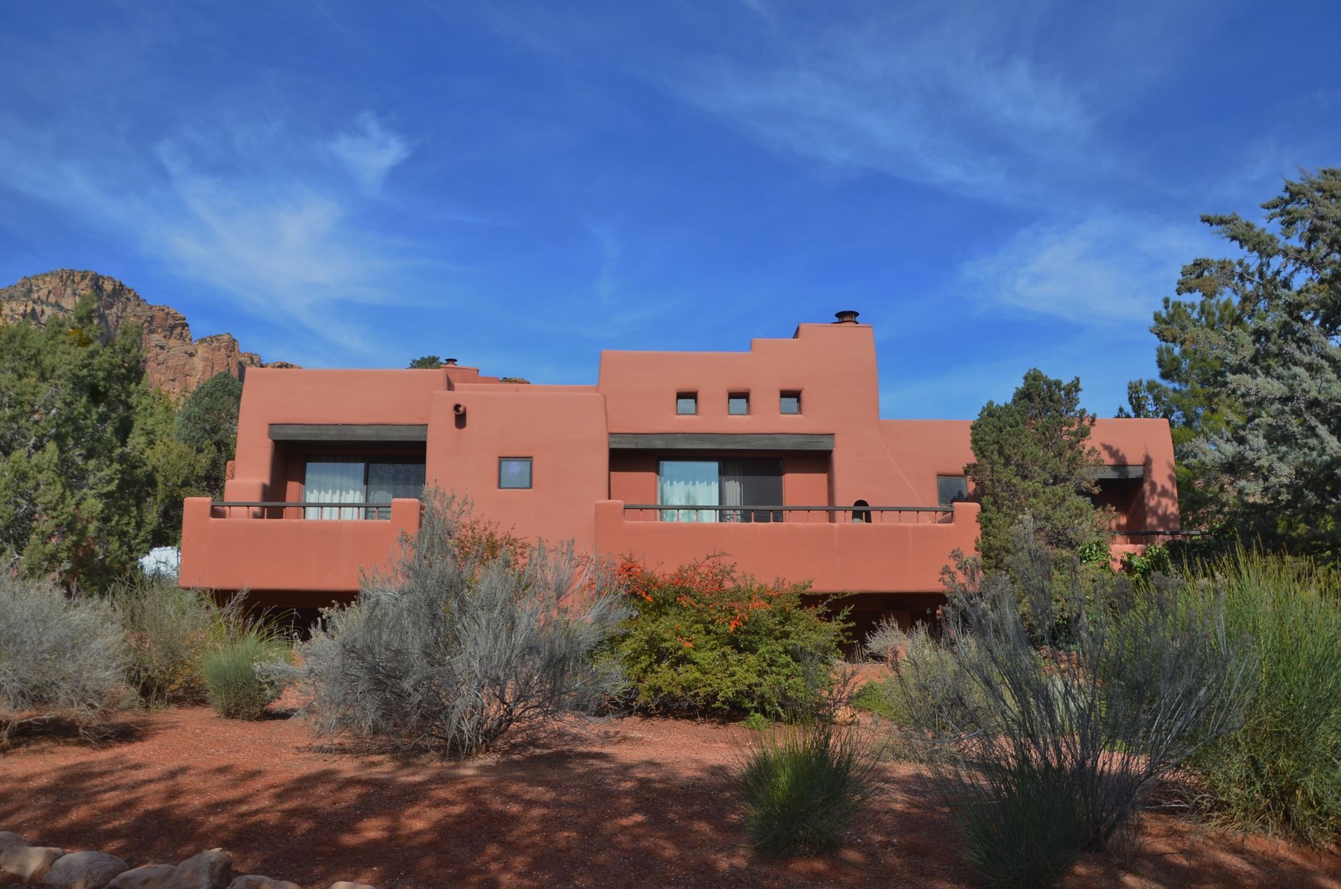 Red stucco building with a flat roof and balcony, against a backdrop of blue sky and red rock formations.