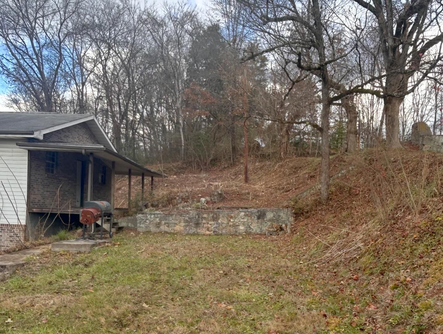 A house with a porch and a small yard. The yard has a retaining wall and is surrounded by trees.