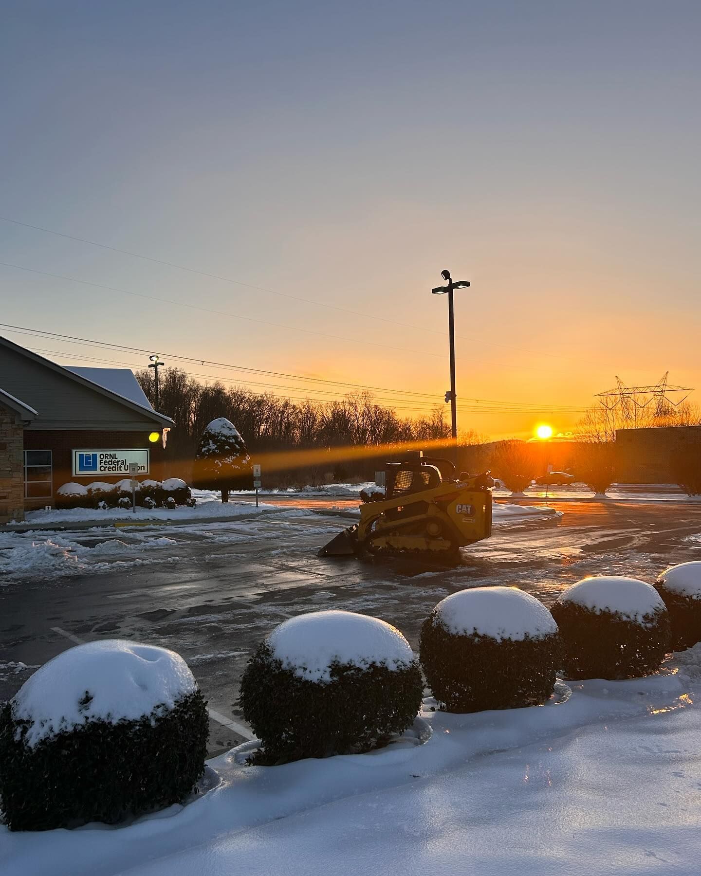 Snowy parking lot at sunrise; small bulldozer; snow-covered shrubs; building on the left.