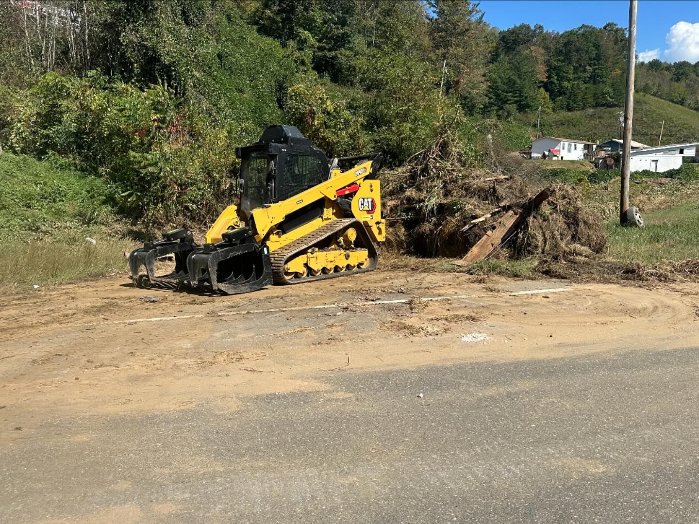 Yellow skid steer operating near a roadside, clearing debris. Green foliage and a power pole are in the background.