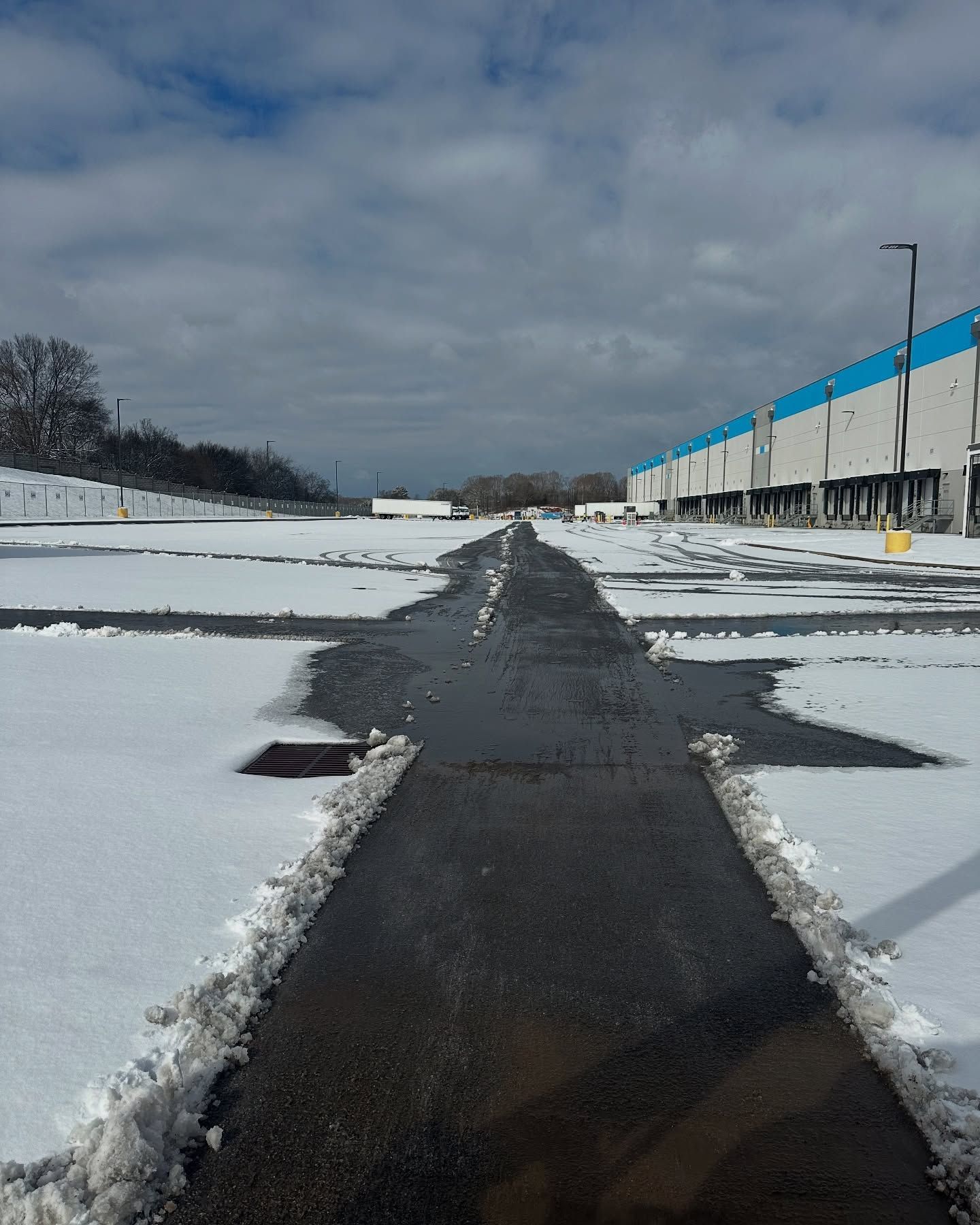 Snowy paved area leading to warehouse, partly melted with snow on the sides, under cloudy sky.