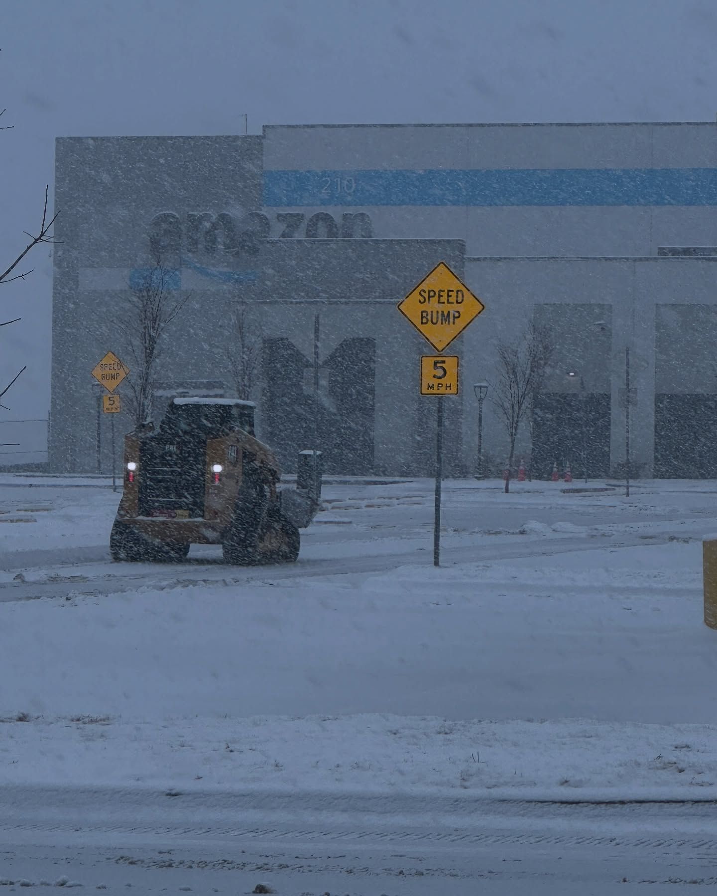 A yellow front-end loader plowing snow in front of an Amazon warehouse during a snowstorm.