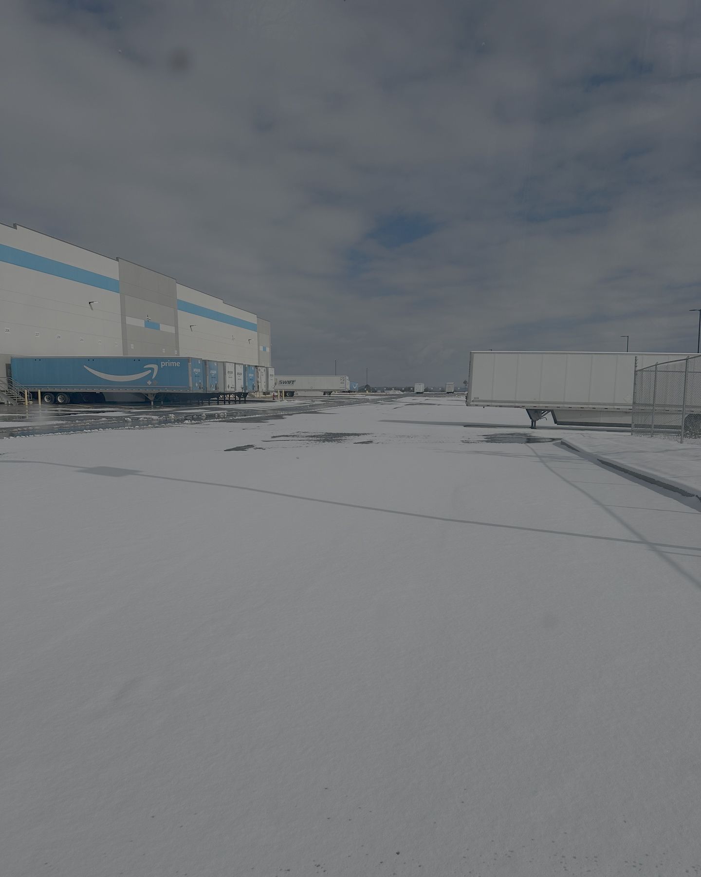 Snow-covered parking lot with an Amazon warehouse and trucks against a cloudy sky.