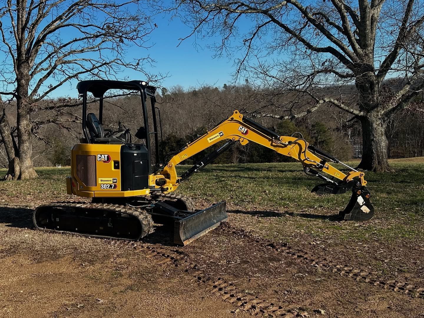 Yellow mini excavator on a dirt patch, under a blue sky, near trees.