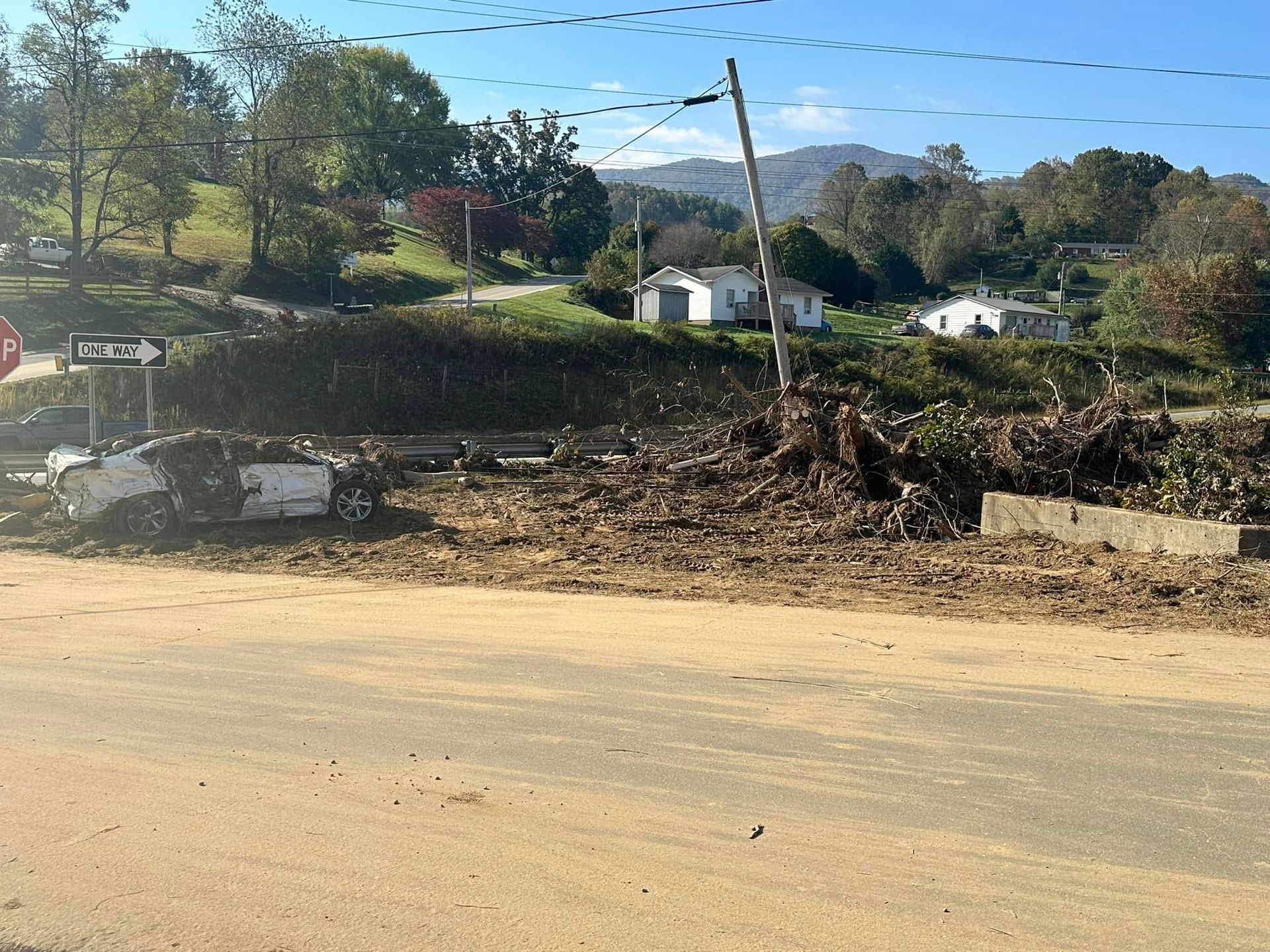 Debris covers a road with a damaged car, near homes and a hillside under a blue sky.