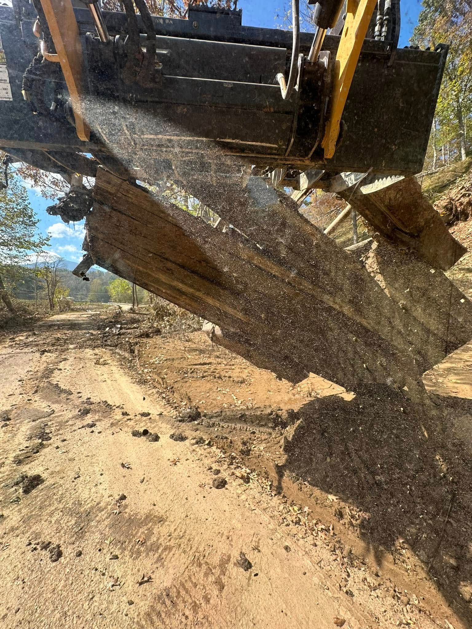 Excavator mulching wood into the ground, creating a rough path in a forest setting.