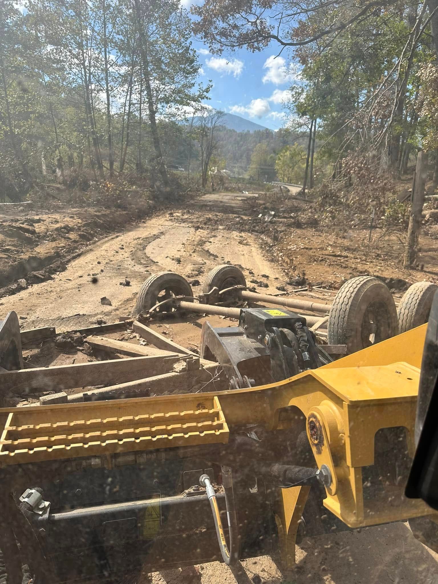 A yellow grading machine working on a dirt road in a wooded area on a sunny day.