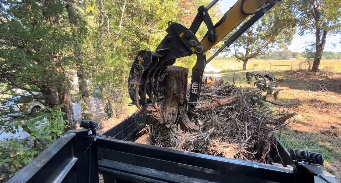 An excavator's claw gripping a tree stump and debris, placing it into a black dump bed. Outdoors, trees in background.