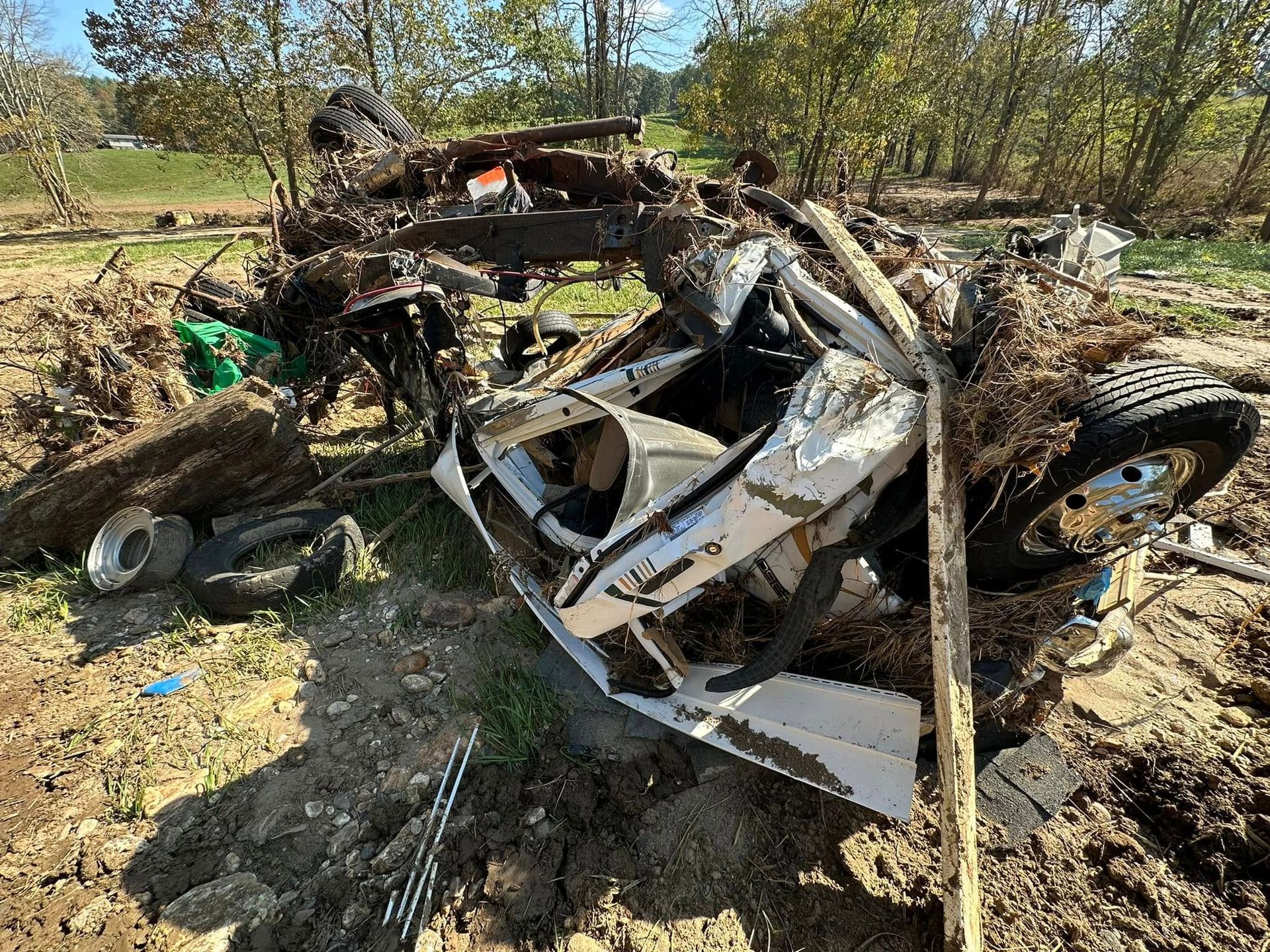 Wrecked white car, heavily damaged, sits on muddy ground, surrounded by debris, near green foliage.