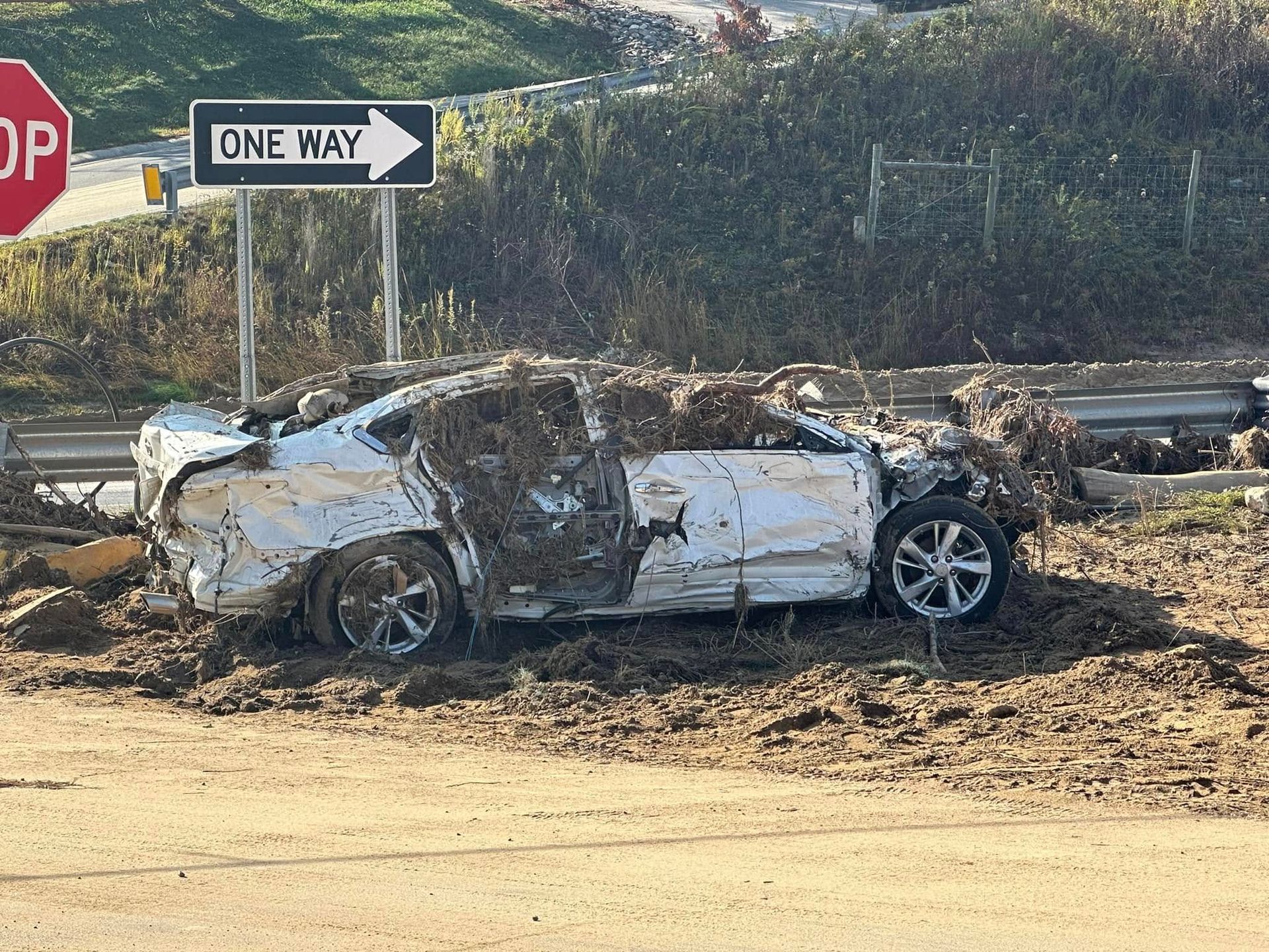 Damaged white car covered in mud and debris, next to a stop sign and one-way sign.