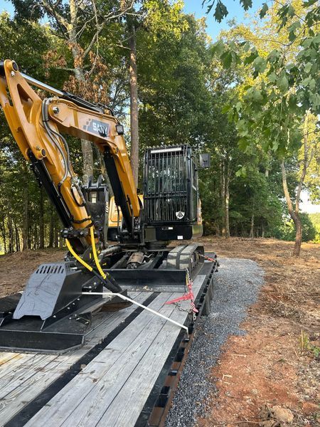 Yellow excavator on a flatbed trailer on a gravel driveway in a wooded area.