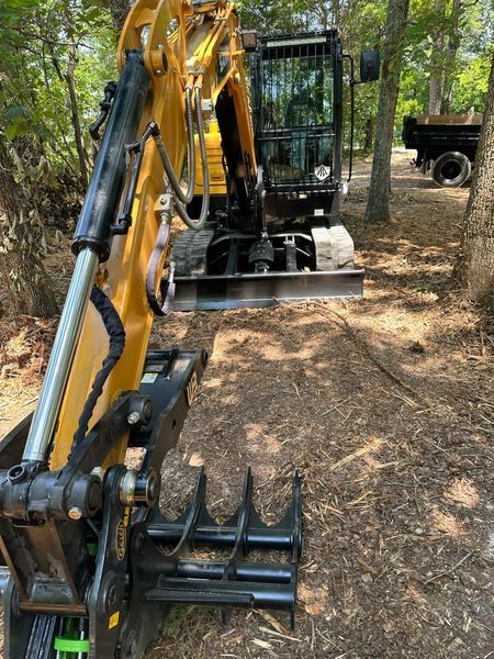 Yellow excavator with a grapple attachment on a dirt path in a wooded area.