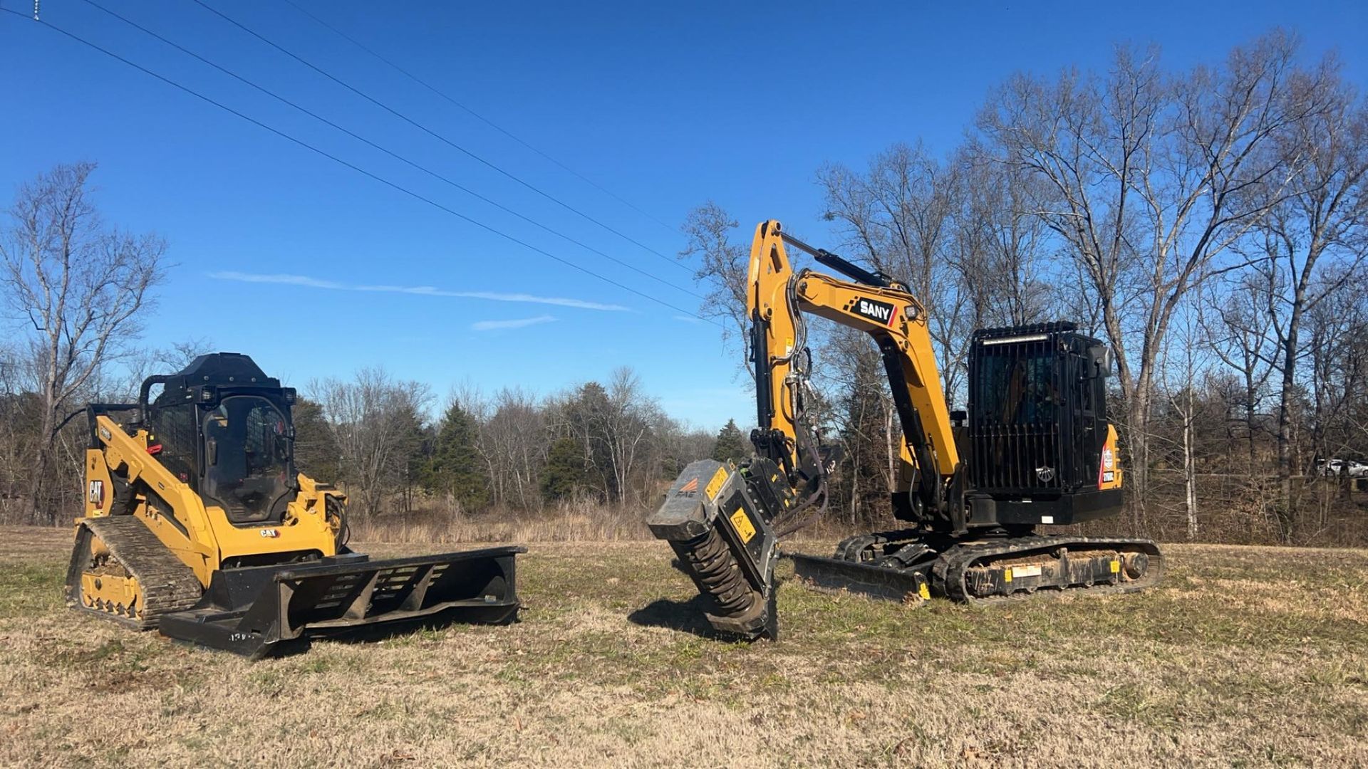 Yellow excavator and skid steer on a grassy field under a blue sky, near trees.