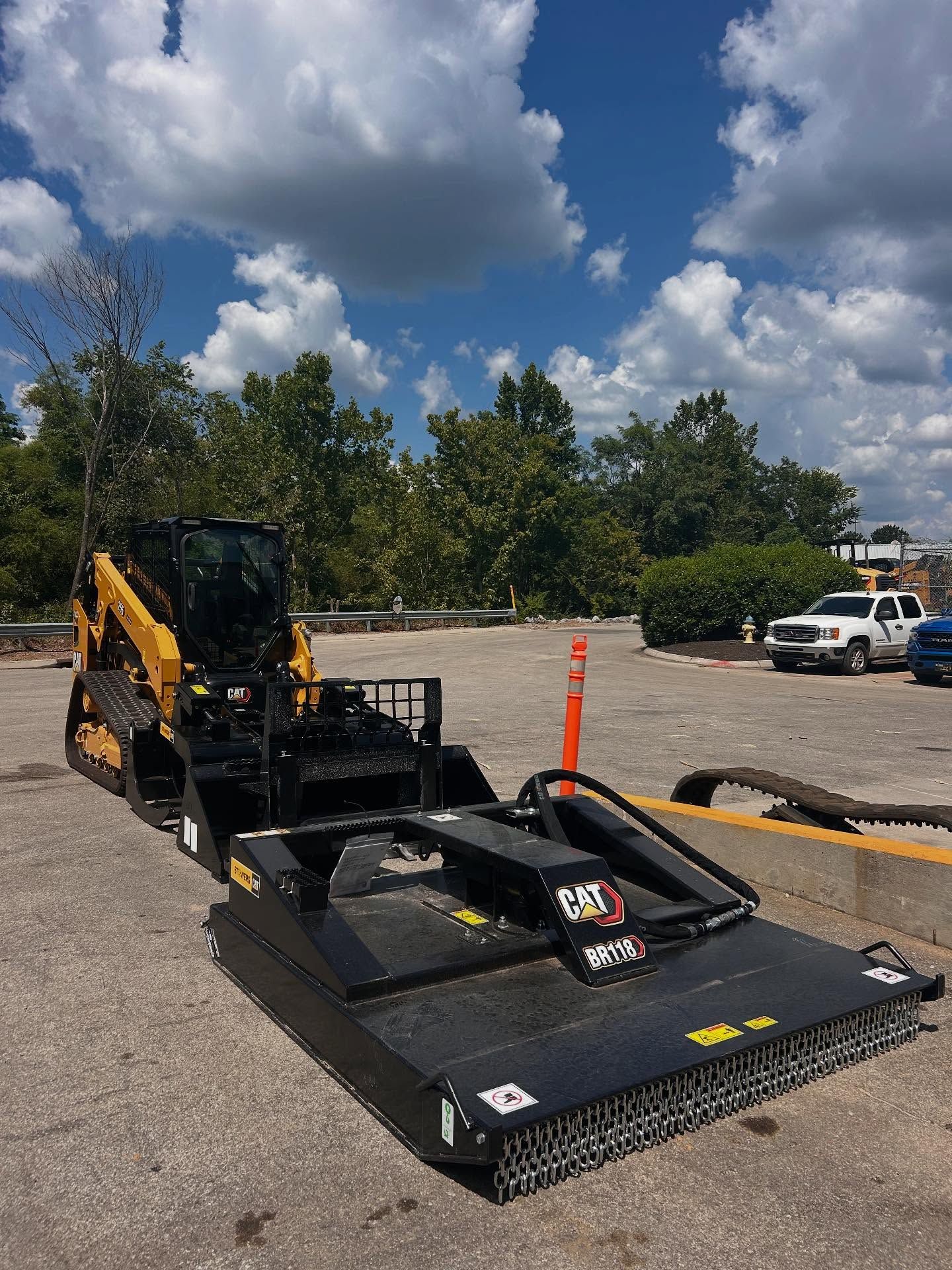 Yellow and black Caterpillar skid steer with brush cutter attachment on pavement, cloudy sky.