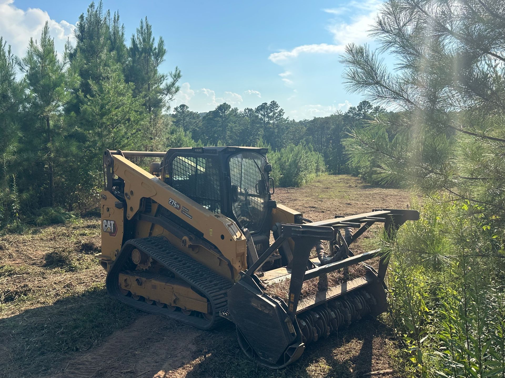 Yellow skid steer with forestry cutter clearing a wooded area under a blue sky.