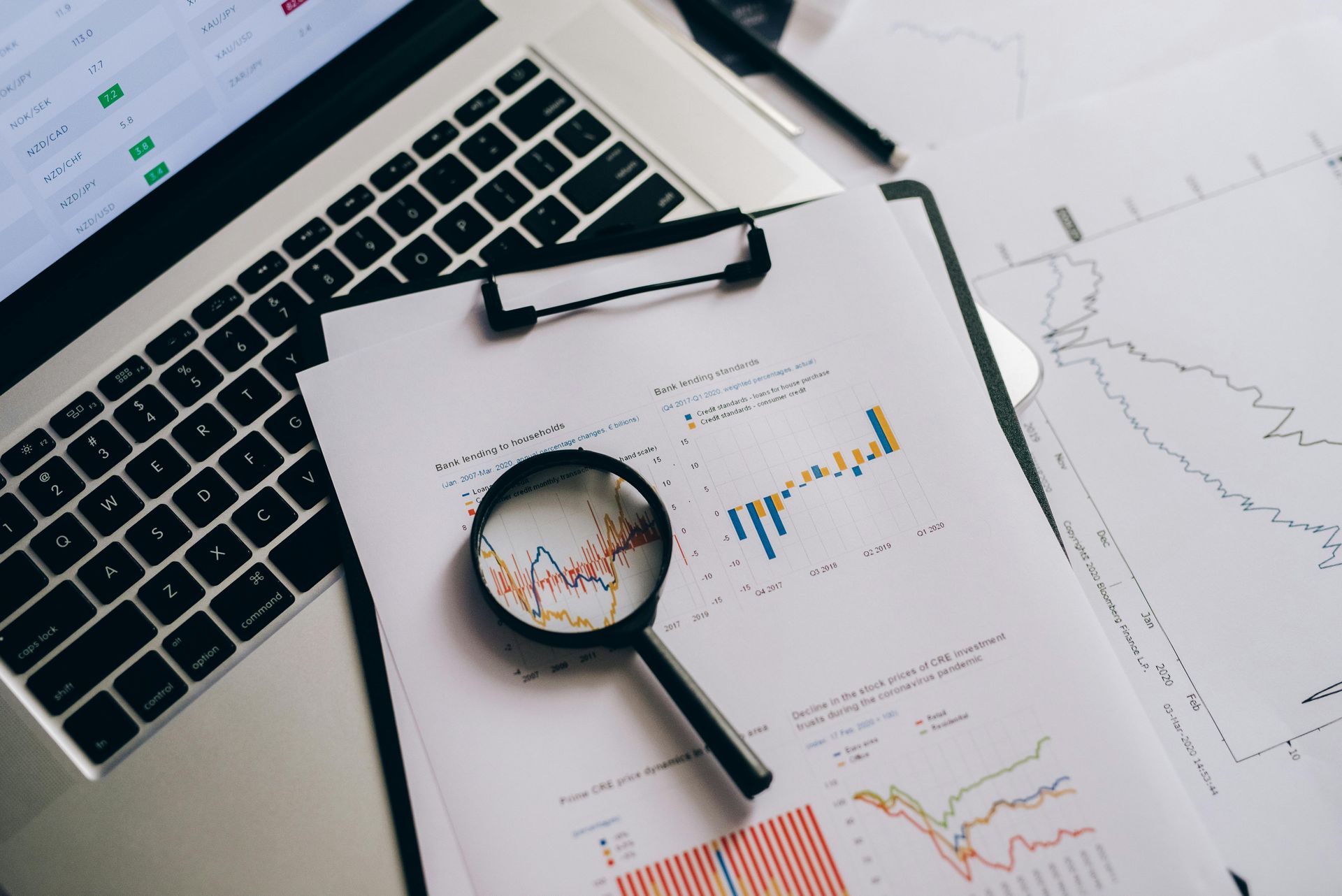 Laptop and clipboard with financial charts and a magnifying glass on a desk