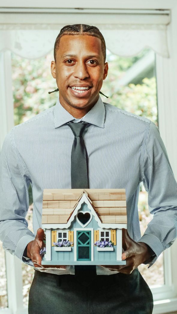 Man holding a small decorative house model in front of a window, wearing a striped shirt and tie.