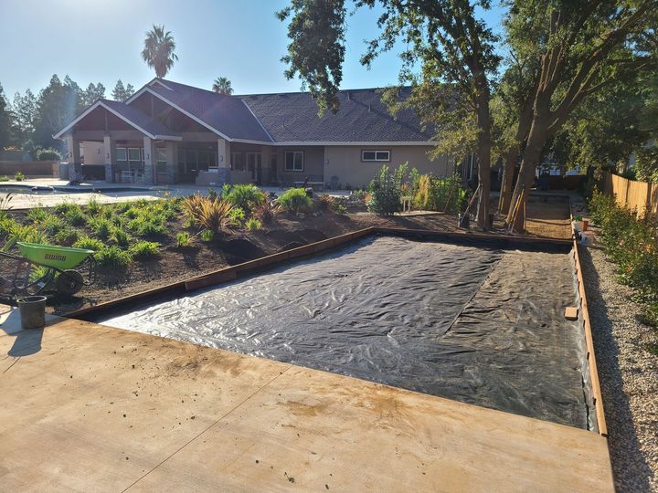 A landscaped front yard with a rectangular dirt lot beside a house and driveway, bordered by trees and shrubs.