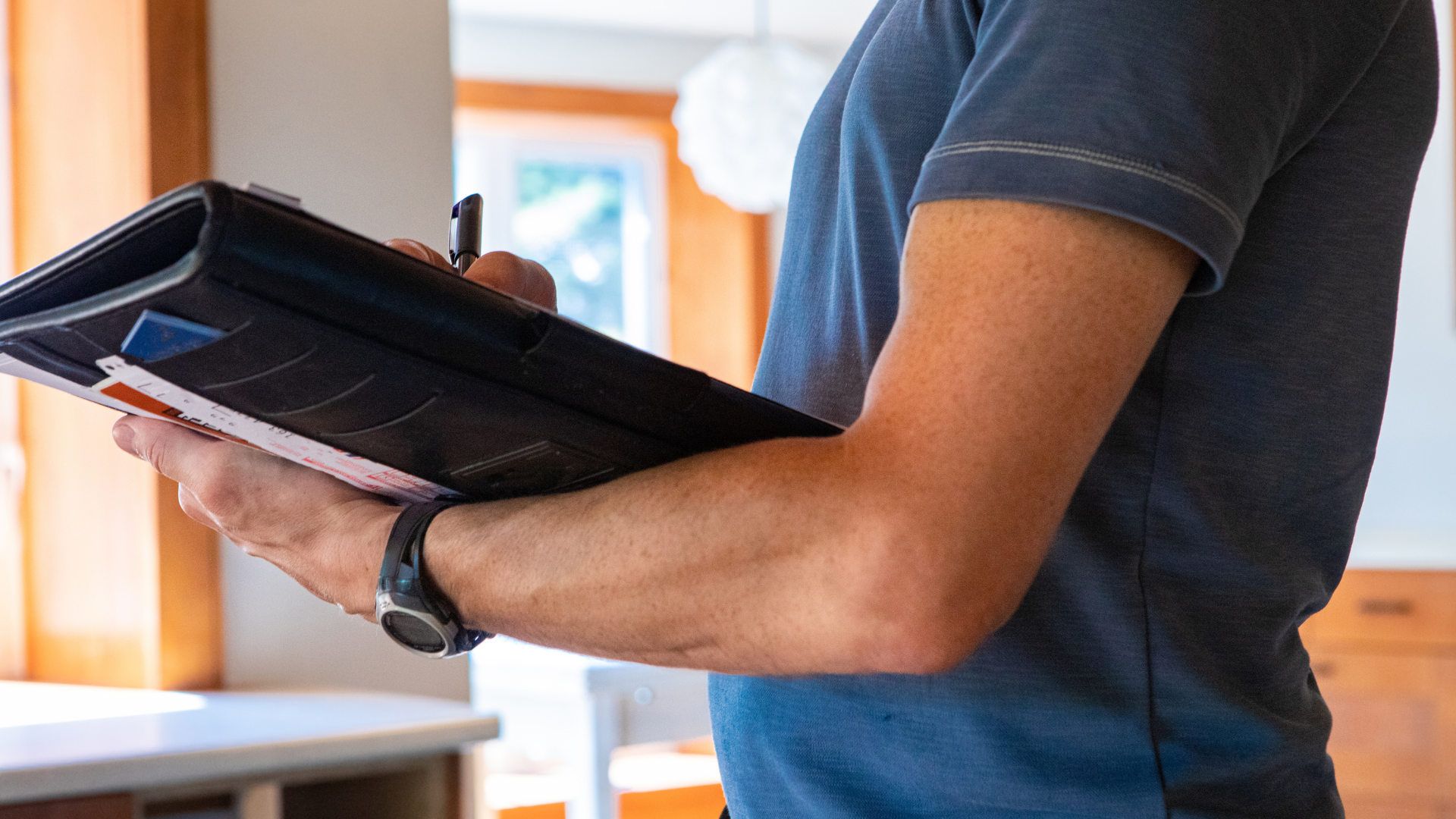 Man in gray shirt writing in a black portfolio, indoors near a window.