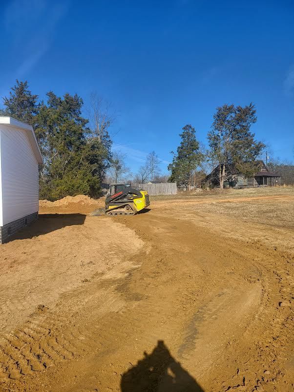 A yellow skid steer moves dirt on a sunny, rural construction site next to a white house.