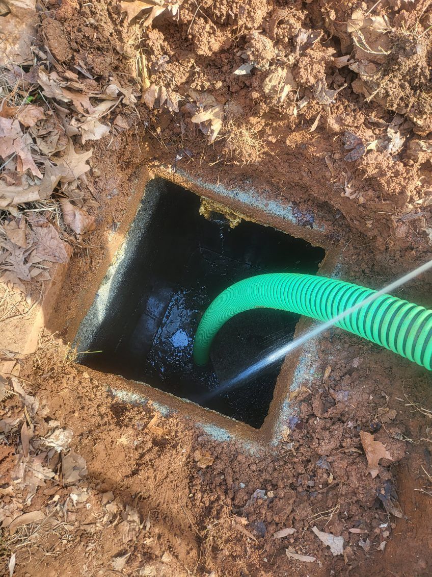 Yellow excavator digging a trench in red dirt, house in background.